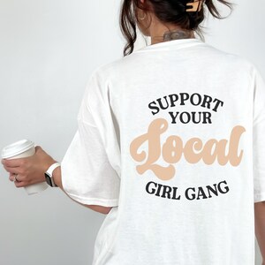 May include: White t-shirt with a black graphic that says "Support Your Local Girl Gang" in a retro font. The word "Local" is in a peach color.