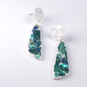 Op de afbeelding: Een paar oorhangers met witte, iriserende en blauwgroene accenten. Elke oorbel heeft een witte, cirkelvormige oorbelknop en een driehoekige hanger met een mozaïek van iriserende stukjes. Op een witte achtergrond.