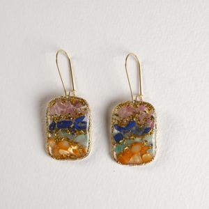 May include: A pair of rectangular earrings with gold-colored frames. Each earring features layers of colorful gemstones, including pink, blue, green, and orange, with gold flecks throughout. The earrings have a simple hook design.