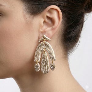 Può includere: Orecchini dorati a dichiarazione con un design strutturato e organico. Gli orecchini presentano una forma ad arco con accenti pendenti a forma di goccia. La superficie ha un motivo bianco e oro e gli orecchini sono attaccati con elementi dorati.