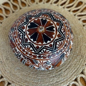 May include: A round, carved gourd with a brown and black geometric design. The gourd has a small, white, round object in the center.