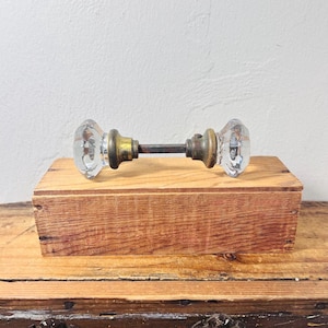 May include: A pair of clear, faceted glass door knobs connected by a central metal spindle. Each knob features a circular brass-toned base plate that attaches to the glass. The hardware is positioned horizontally, showcasing the reflective glass.