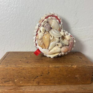 May include: A heart-shaped decorative object made of various seashells, with a red ribbon accent. The shells are in shades of white, beige, and tan. The object is resting on a wooden surface.