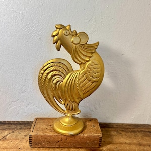 May include: A gold-colored rooster figurine with detailed molded feathers, a prominent comb, and a circular base. The rooster is mounted on a rectangular, unfinished wooden block that serves as a stand for the metallic decorative object.
