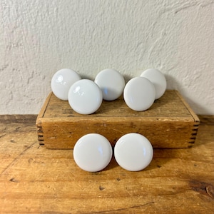 May include: A set of six round, smooth, white ceramic knobs. Each knob features a simple, circular design with a glossy finish. The knobs are arranged with four positioned on top of a rectangular wooden block and two placed in front of the block.
