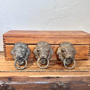 May include: Three matching metal lion head door knockers with a weathered bronze finish. Each piece features a detailed lion face with a mane and a circular ring held in the lion's mouth. The knockers are mounted on a rectangular wooden box.