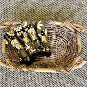 May include: A woven wicker serving tray with handles holds four rolled floral napkins secured by gold-toned napkin rings. The napkins feature a black, olive green, and cream floral pattern. The tray sits on a textured, neutral-toned surface.