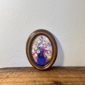 May include: An oval-shaped wall art piece featuring a floral arrangement in a deep blue vase. The bouquet includes various colorful flowers in shades of pink, white, yellow, and purple. The artwork is encased in a dark brown wooden frame.