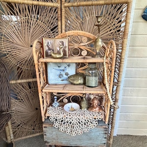 May include: A vintage rattan shelf unit with three shelves, displaying various antique items. Includes a small framed picture, a brass candlestick, a glass jar, and decorative objects. The unit is set against a woven screen.