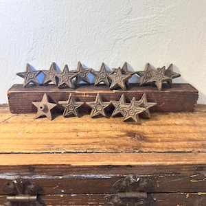 May include: A collection of small, five-pointed star-shaped metal objects with a dark, weathered finish. Each star features a central circular detail. Several stars are arranged in a row atop a rectangular wooden block, while others are placed in front.