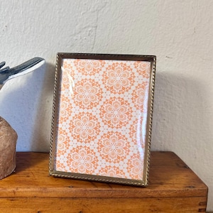 May include: A rectangular picture frame with a metallic, twisted rope-style border. The frame contains a decorative insert featuring a repeating white floral lace pattern set against a vibrant orange background, protected by a clear glass pane.