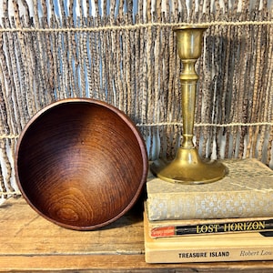 May include: A wooden bowl with a dark brown finish sits next to a brass candlestick holder. The candlestick is on top of a stack of vintage books, including "Lost Horizon" and "Treasure Island."