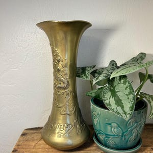 May include: A tall, brass-colored vase with a flared top and intricate dragon and floral relief carvings. The vase is displayed next to a teal ceramic pot with a plant. The vase is on a wooden surface.