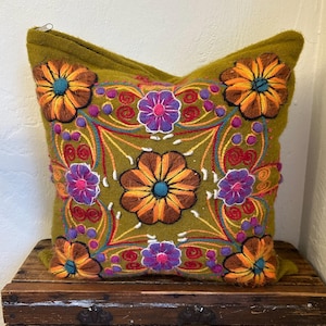 May include: Olive green square pillow with embroidered floral design. The pillow features large flowers in shades of orange, brown, and purple, with blue centers. The embroidery includes red, yellow, and white details, creating a vibrant, decorative accent.