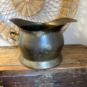 May include: Antique brass pitcher with a wide spout and a small handle. The pitcher has a rounded body and sits on a circular base. The brass has a tarnished, aged appearance, suggesting it is vintage. The background includes a woven wall hanging and a wooden surface.