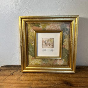 May include: A square gold-toned frame containing a small central illustration of a bridge and buildings with the text "BRUGGE" and "begijnhof" below it. The inner border features a floral pattern with muted pink, green, and yellow tones.