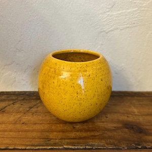 May include: A round, yellow ceramic vase with a speckled pattern. The vase has a wide opening and sits on a wooden surface. The interior of the vase is a darker yellow.