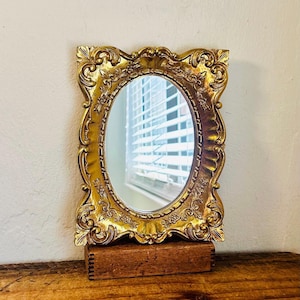 May include: An oval mirror with a gold-colored frame featuring ornate, raised scrollwork and floral detailing. The mirror is mounted on a rectangular, dark brown wooden base with visible finger joints on the sides.