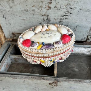 May include: An oval-shaped jewelry box adorned with seashells, red, white, and purple shells, and small colorful beads. The box has a cream-colored base and a lid with the word "Clubagne" written on it.
