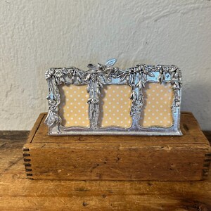 May include: A silver-toned metal triple picture frame featuring a decorative relief of hanging floral garlands and leaves. The three rectangular openings contain inserts with a light yellow background and a repeating pattern of small white dots.
