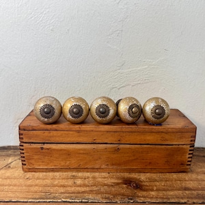 May include: Five spherical drawer knobs with a distressed gold finish and a dark bronze-toned metal center rosette. Each knob features a textured, weathered surface and is arranged in a horizontal row on a rectangular wooden box.