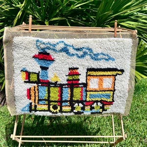 May include: A rectangular punch needle embroidery piece featuring a colorful steam train design on a white background. The train is rendered in shades of blue, red, yellow, and orange with black outlines, and includes a plume of blue smoke above.