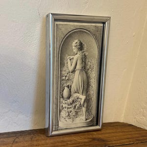 May include: A rectangular wall plaque featuring a monochromatic relief sculpture of a figure standing beside a water pitcher and tall grasses, enclosed within an arched frame. The artwork is encased in a polished silver-toned metal frame.