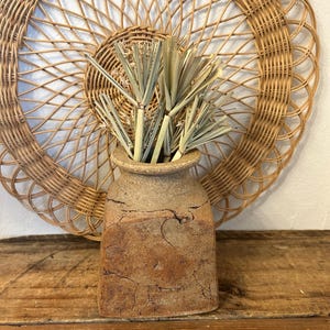 May include: A rustic, textured, brown ceramic vase with a square base and a round opening, filled with dried, light green grass. A large, woven, circular wall hanging is in the background, creating a natural, earthy aesthetic.