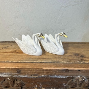 May include: Two white ceramic swan figurines with detailed feather textures. The swans have black eyes and yellow beaks. They are displayed on a wooden surface, showcasing a classic decorative item.