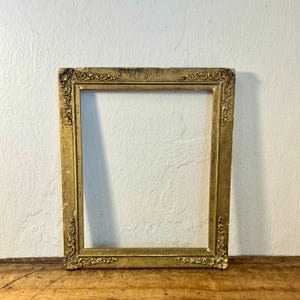 May include: A rectangular picture frame with a distressed gold finish. The frame features ornate, raised scrollwork and floral detailing at each of the four corners. The surface shows visible signs of wear and aging, revealing darker tones beneath.