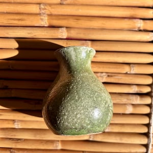 May include: A small, bulbous ceramic vase with a textured, mottled green glaze. The vessel features a narrow neck that flares slightly at the rim, with a rounded body that tapers toward the base, showcasing a speckled finish throughout the surface.