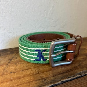 May include: Green and white striped belt with a brown leather interior and a silver buckle. The belt has a navy blue embroidered "K" and is rolled up on a wooden surface.
