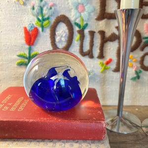 May include: A clear glass paperweight with a vibrant blue abstract design sits atop a stack of vintage books. A silver candle holder with a white candle is to the right. The background features a white embroidered wall hanging with colorful floral designs.