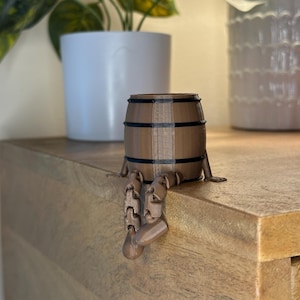 Puede incluir: Una maceta marrón en forma de barril con bandas negras y patas articuladas. La maceta está sobre una superficie de madera, con dos macetas blancas en el fondo.