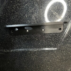 May include: A black plastic mounting bracket with four screw holes. The bracket is attached to a black surface with a speckled pattern.