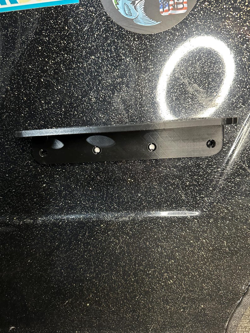 May include: A black plastic mounting bracket with four screw holes. The bracket is attached to a black surface with a speckled pattern.