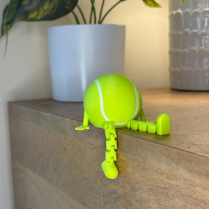 May include: A neon green tennis ball figurine with white stripes and articulated legs and feet. The figurine is sitting on a wooden surface, with a white potted plant in the background.
