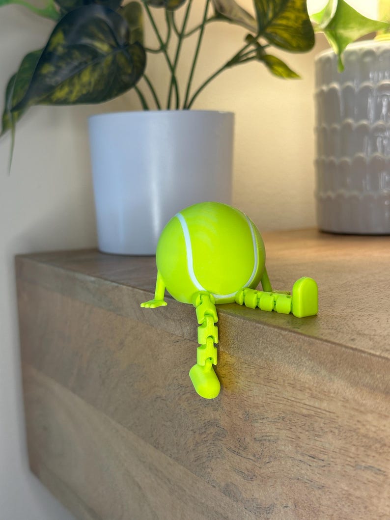 May include: A neon green tennis ball figurine with white stripes and articulated legs and feet. The figurine is sitting on a wooden surface, with a white potted plant in the background.
