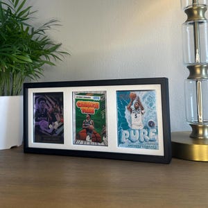 May include: A black framed display featuring three basketball trading cards. The cards showcase Anthony Edwards, with text including "Crunch Time" and "Pure". The frame is on a wooden surface, with a lamp and plant in the background.