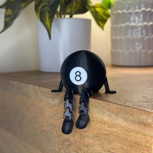 Könnte beinhalten: Eine schwarze 8-Ball-Figur mit beweglichen Beinen und kleinen Füßen. Die Zahl 8 ist in einem weißen Kreis auf dem Ball dargestellt. Die Figur sitzt auf einer Holzoberfläche.