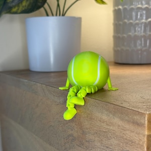 May include: A whimsical, lime green tennis ball figurine with white stripes and articulated legs and arms. The figurine is posed as if sitting on a wooden surface. Decorative plants in white pots are in the background.