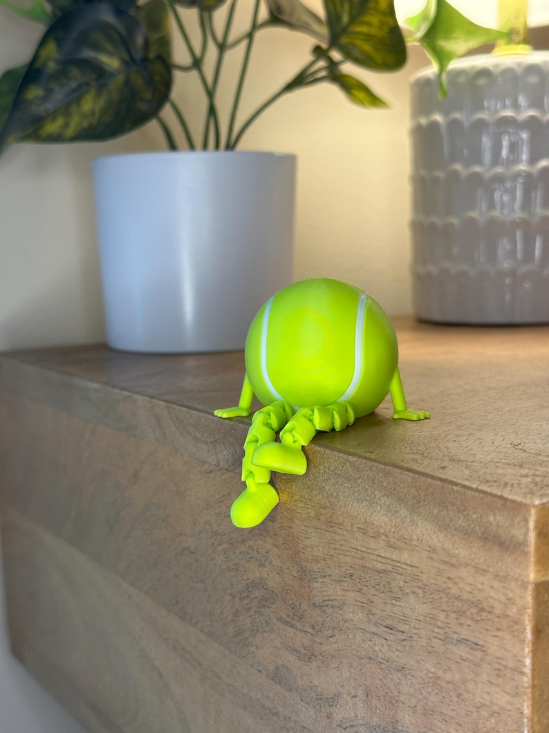 May include: A whimsical, lime green tennis ball figurine with white stripes and articulated legs and arms. The figurine is posed as if sitting on a wooden surface. Decorative plants in white pots are in the background.