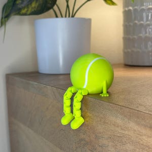 May include: A whimsical, neon green tennis ball figurine with articulated legs and feet, sitting on a wooden surface. The tennis ball has a white stripe and is positioned near a white potted plant. The figurine is a playful decorative item.