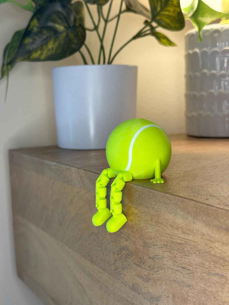 May include: A whimsical, neon green tennis ball figurine with articulated legs and feet, sitting on a wooden surface. The tennis ball has a white stripe and is positioned near a white potted plant. The figurine is a playful decorative item.