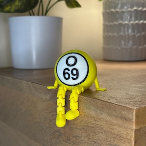 Pode incluir: Uma bola de bingo amarela com o número 69 impresso em um círculo branco. A bola tem pequenos braços e pernas articuladas, dando-lhe uma aparência lúdica. O objeto está em uma superfície de madeira.