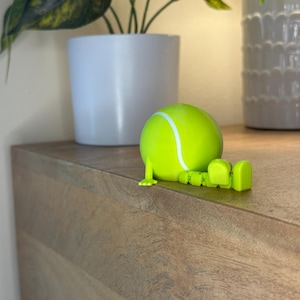 May include: A lime green tennis ball figurine with articulated legs and feet, resting on a wooden surface. The tennis ball has a white stripe and is positioned near a white potted plant. The background includes another white pot and a green plant.