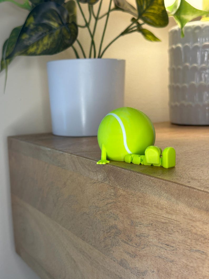 May include: A lime green tennis ball figurine with articulated legs and feet, resting on a wooden surface. The tennis ball has a white stripe and is positioned near a white potted plant. The background includes another white pot and a green plant.