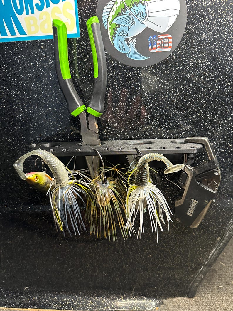 May include: A black fishing lure organizer with three white, green, and black fishing lures hanging from it. A pair of green and black pliers are hanging from the top of the organizer. A black tool with the word "Rampage" printed on it is hanging on the right side of the organizer.
