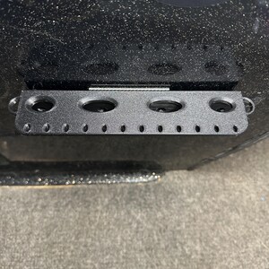 May include: Black metal rod with five round holes and a row of smaller holes. The rod is mounted on a black surface.
