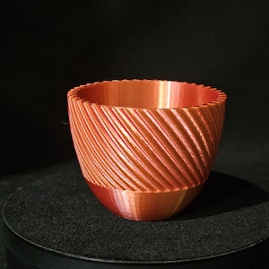 Può includere: Una ciotola color rame, stampata in 3D, con un motivo a righe diagonali strutturate. La ciotola ha una base arrotondata e un bordo scanalato. L'oggetto è esposto su una superficie nera su sfondo scuro.
